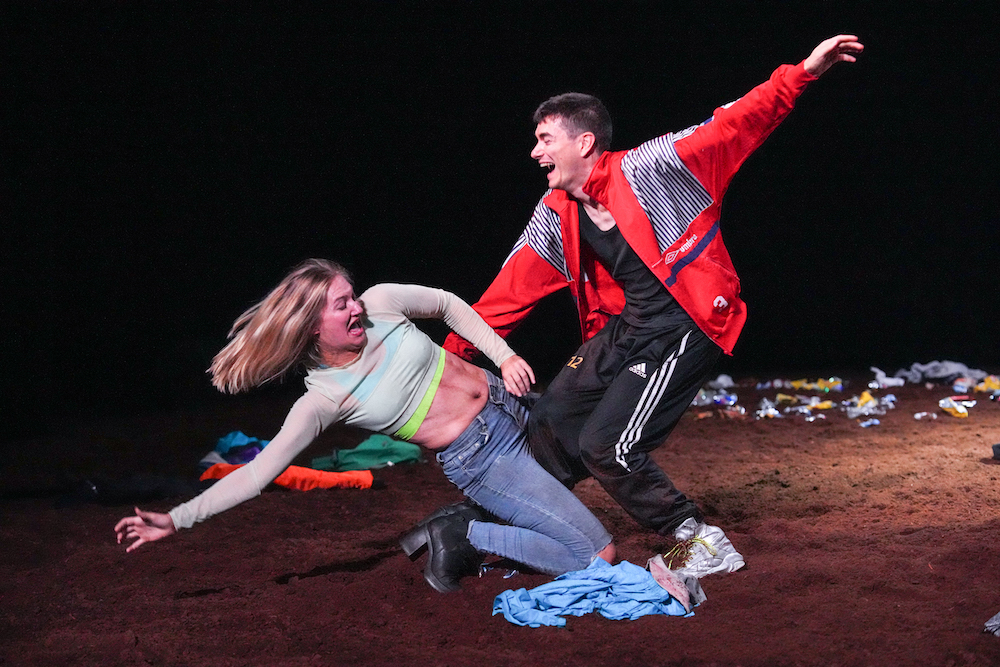 two people laughing so hard that one falls backwards and the other tries to pick her up. She is in a midriff top and jeans, he is in a red jacket wearing black and white sports pants.  